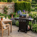 Mid Ulster Garden Centre Broil King Crown 310 Shadow matte black gas BBQ on a patio in a lush garden setting.